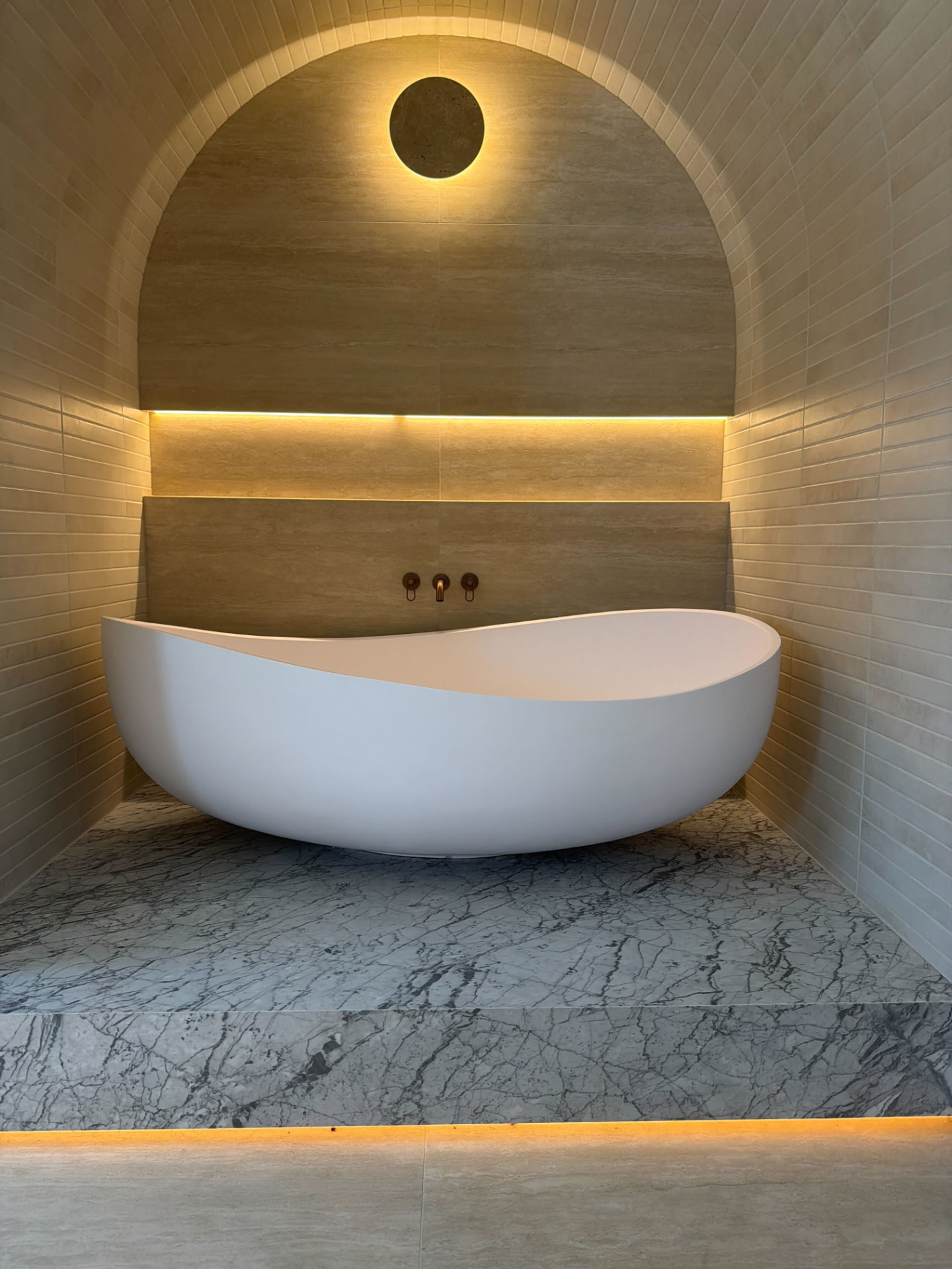 Arched bathroom alcove with warm LED strip lighting and a feature wall light above a freestanding bath
